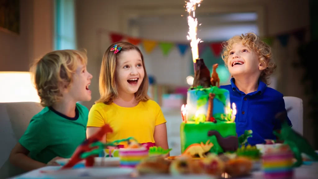 "Children excitedly celebrating a birthday with a dinosaur-themed cake and a sparkling candle, a great inspiration for planning children's parties with fun themes."