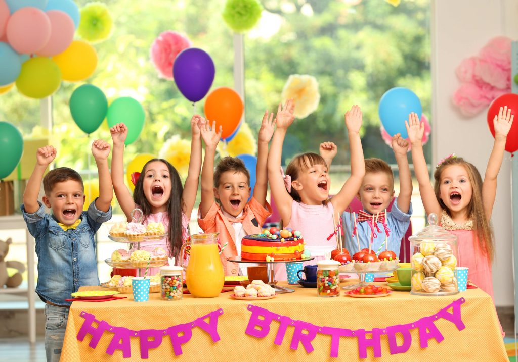 Excited children celebrating a birthday with colorful balloons, a vibrant cake and a table filled with treats set against a bright festive background