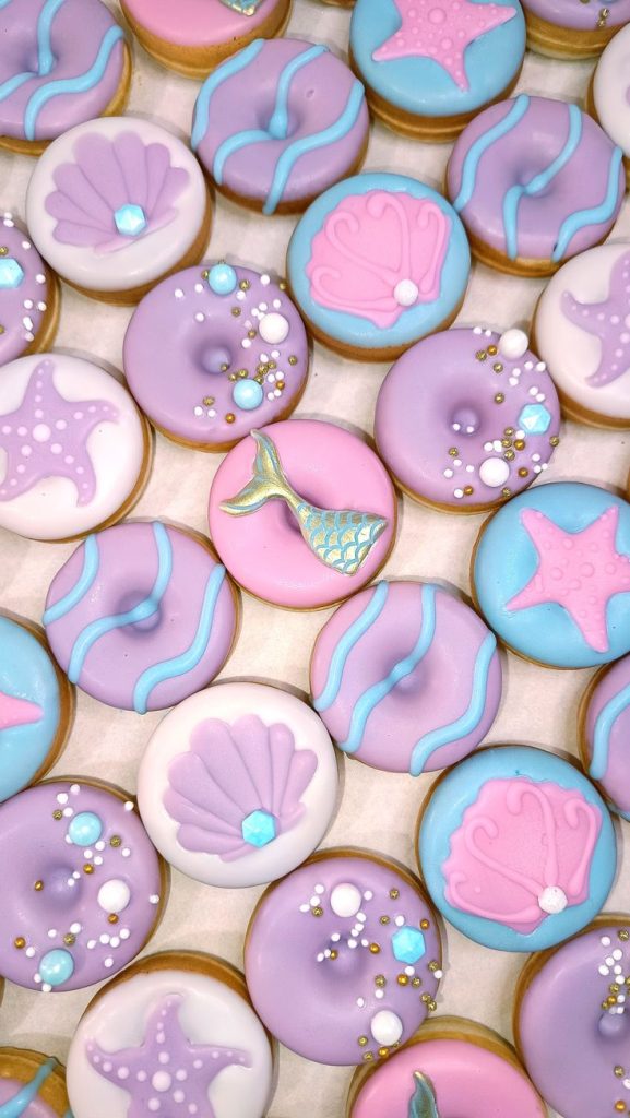 "An assortment of beautifully decorated mini donuts with a mermaid theme. The donuts are adorned in pastel hues of pink, purple, and blue, featuring intricate designs such as seashells, starfish, and mermaid tails. Embellished with edible pearls, gold accents, and wave patterns, these treats exude whimsical under-the-sea vibes, perfect for a magical celebration."