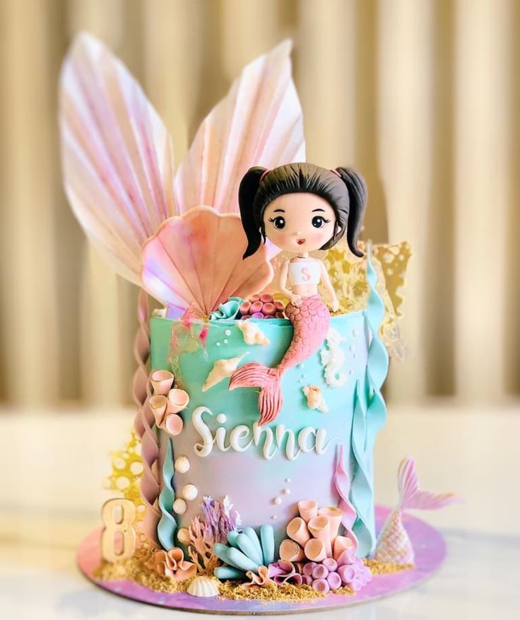 A pastel mermaid-themed cake featuring a 3D topper of a mermaid girl with pigtails, oversized shimmering fins, and intricate sea-inspired decorations like coral and seashells. The gradient of pastel colors adds a magical and playful touch.