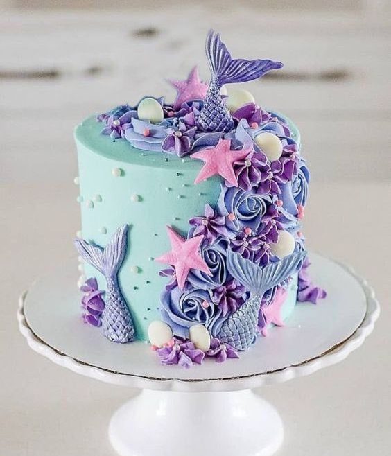 A whimsical turquoise cake adorned with cascading purple buttercream flowers, mermaid tails, starfish, and seashells. It captures a serene under-the-sea theme with delicate details and a soft oceanic palette.