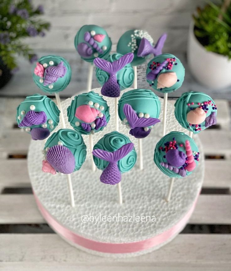 Mermaid-themed cake pops decorated with teal and purple frosting, adorned with edible mermaid tails, seashells, pearls, and ocean-inspired accents, displayed on a white foam stand.