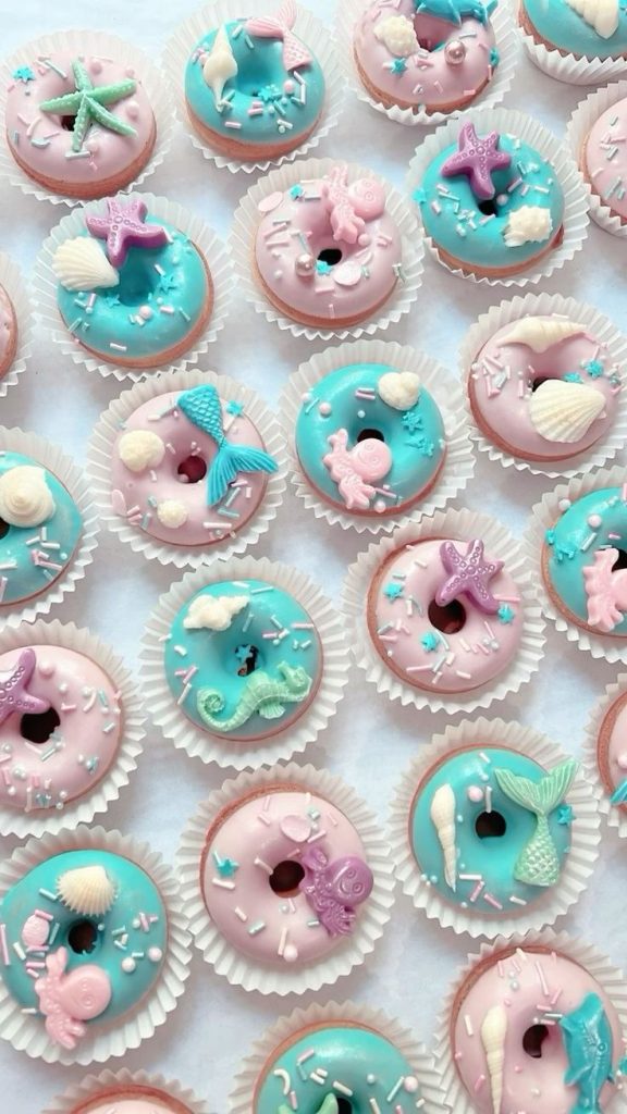 "An elegant display of mini donuts with a mermaid and ocean-inspired theme, featuring pastel pink and blue frosting. Each donut is topped with intricate fondant decorations such as mermaid tails, starfish, seashells, seahorses, and octopuses. Edible sprinkles and pearls add a whimsical touch, making them perfect for a magical under-the-sea celebration."