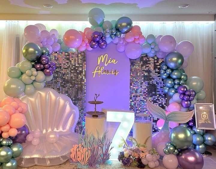 A dreamy under the sea themed balloon backdrop with pastel tones (pink, purple, blue, and green) arranged in clusters. The central focus includes a shiny sequin wall with the your child's name in illuminated script, a large seashell chair, a light-up number 7, and decorative mermaid elements like a tail and underwater plants.