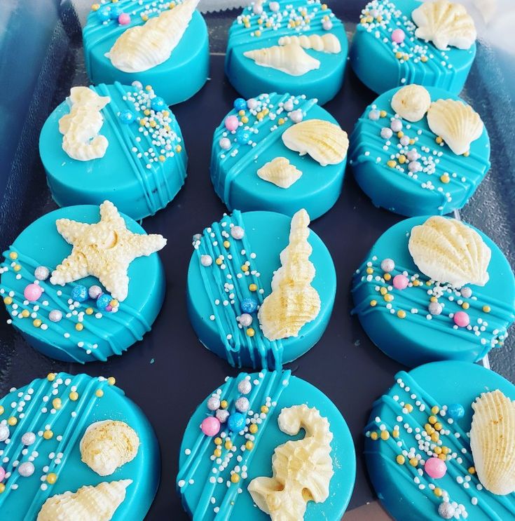 These are beautifully decorated Oreos with an ocean theme. Each piece is covered in blue icing and adorned with edible decorations shaped like seashells and starfish. They are also embellished with colorful sprinkles, adding a festive touch. These treats would be perfect for a under-the-sea themed party or event.