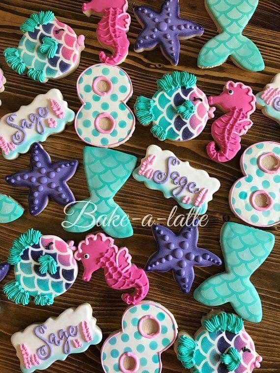 "A vibrant assortment of mermaid-themed sugar cookies in whimsical shapes such as tails, seahorses, starfish, and scallop designs. Decorated in pastel hues of turquoise, purple, and pink, some cookies feature personalized names and numbers, making them ideal for under-the-sea birthday celebrations or ocean-inspired parties."