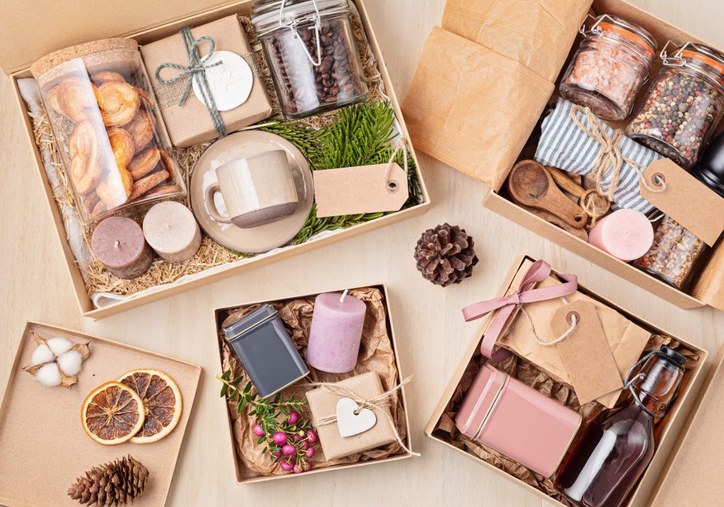 An arrangement of eco-friendly gift boxes on a light wooden surface. Each box contains a variety of items, such as glass jars filled with coffee beans, spices, and salts; a ceramic mug with a saucer; pastel-colored candles; dried fruits; wooden utensils; and small decorative items like heart-shaped ornaments and pinecones. The packaging includes natural fillers like straw and evergreen sprigs, giving the display a rustic, handmade aesthetic. Tags and ribbons complete the minimalist and sustainable presentation.