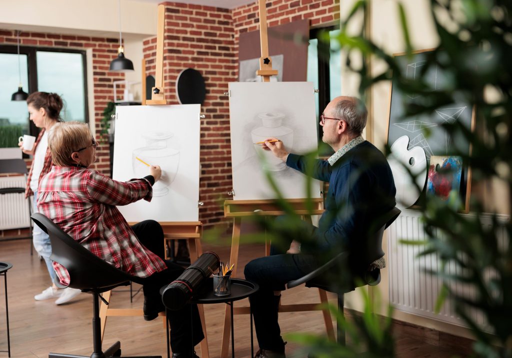 A couple sitting on chairs and sketching still life on easels in an art studio with a cozy brick wall background. The room is bright and filled with natural light, plants, and creative energy