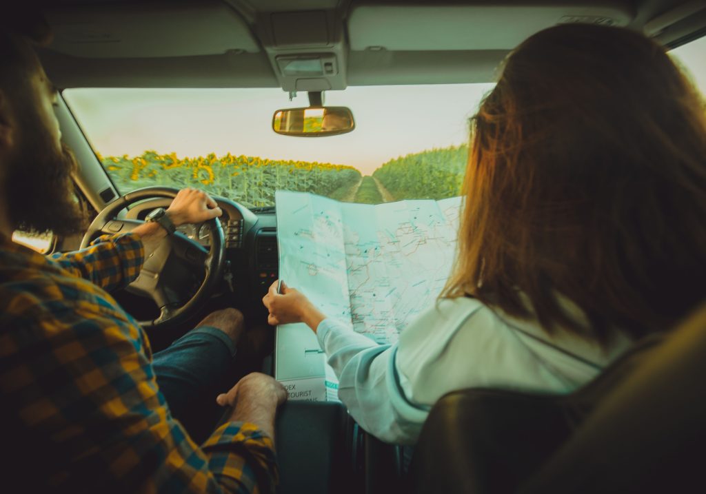 A scenic photo capturing a road trip moment from the car's interior. The driver, wearing a plaid shirt, is holding the steering wheel while the passenger, with long hair, examines a detailed map. Outside, fields of blooming sunflowers stretch along a dirt road under the golden glow of the evening sun, creating a warm and adventurous atmosphere.
