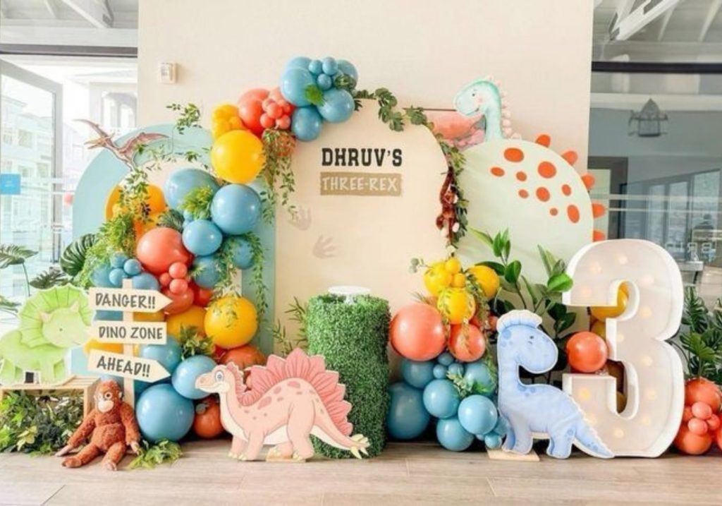 A vibrant dinosaur-themed party backdrop with colorful balloons, greenery, and dinosaur cutouts featuring a light-up number "3" and playful signage.