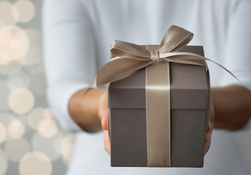 Alt Text: A close-up of hands presenting a gift box wrapped in elegant gray paper with a beige satin ribbon tied in a bow, set against a soft, blurred background with warm bokeh lights.